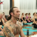 A bearded male student seated between other students during a heated 26 and 2 class at the YogaFX studio